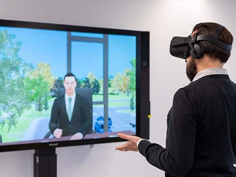 Jan Fiedler trägt eine Virtual Reality Brille, steht vor einem Monitor, auf dem KI-gestützte Avatare zu sehen sind